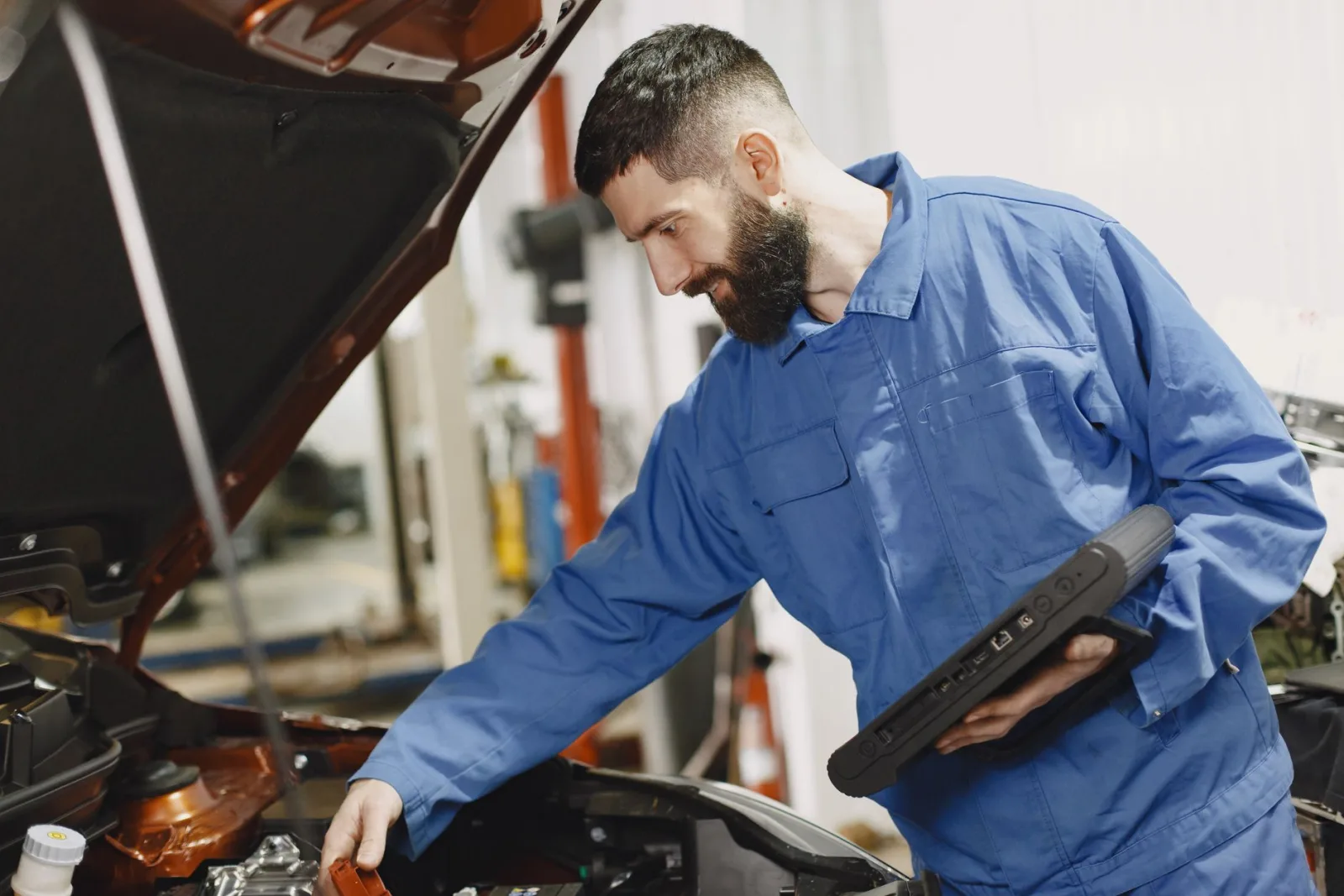 An auto mechanic in blue coveralls inspecting a car's engine in a garage.