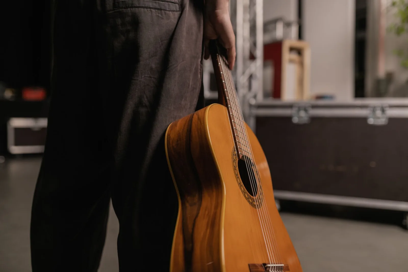 A person holding an acoustic guitar with a backstage setup in view, conveying a musical theme.