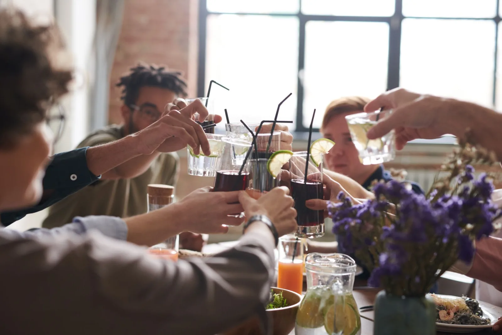 A group of friends toasting drinks in a warm, cozy indoor setting with food and flowers.