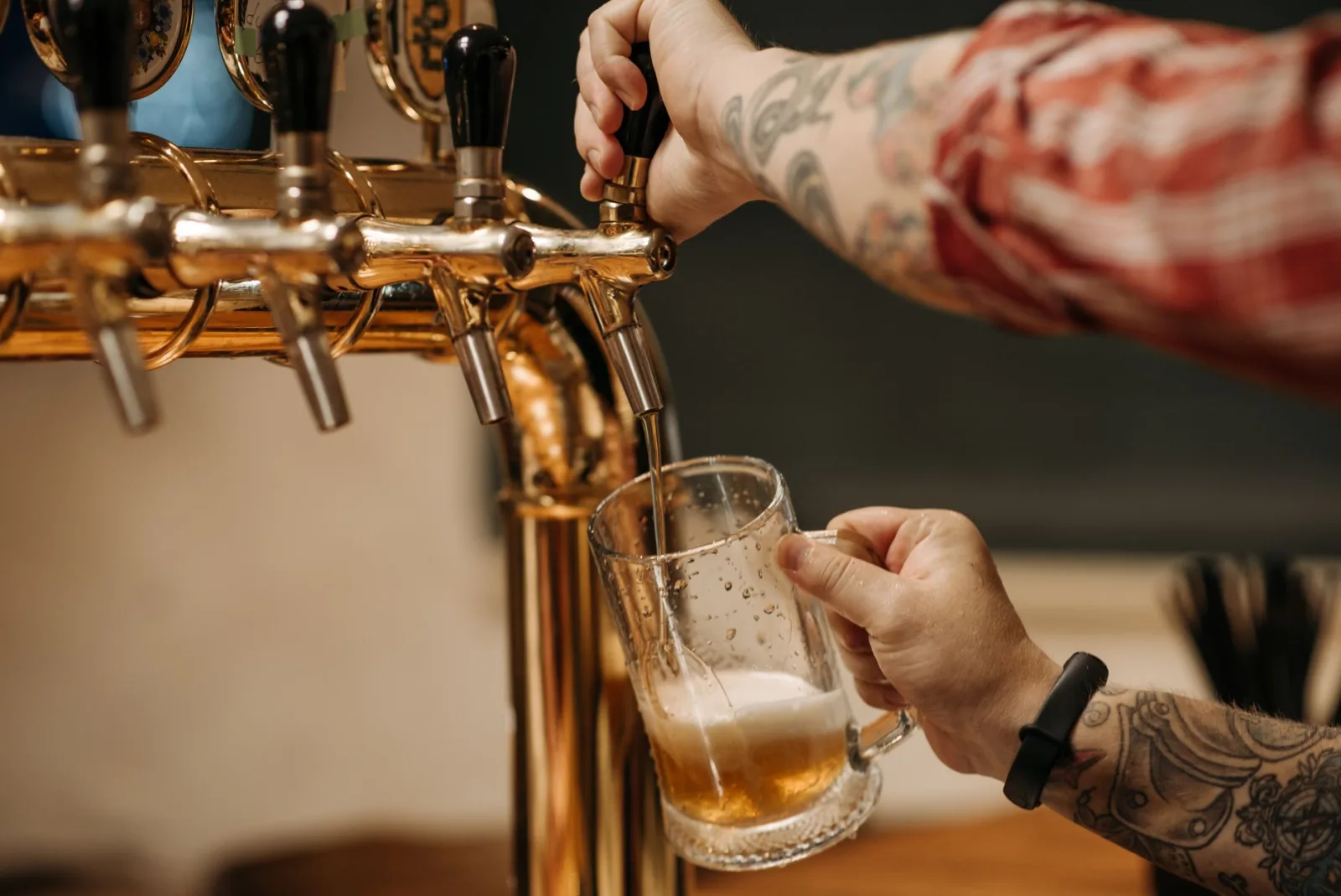 Tattooed bartender pouring a pint of beer from a golden tap, close-up view at a bar.
