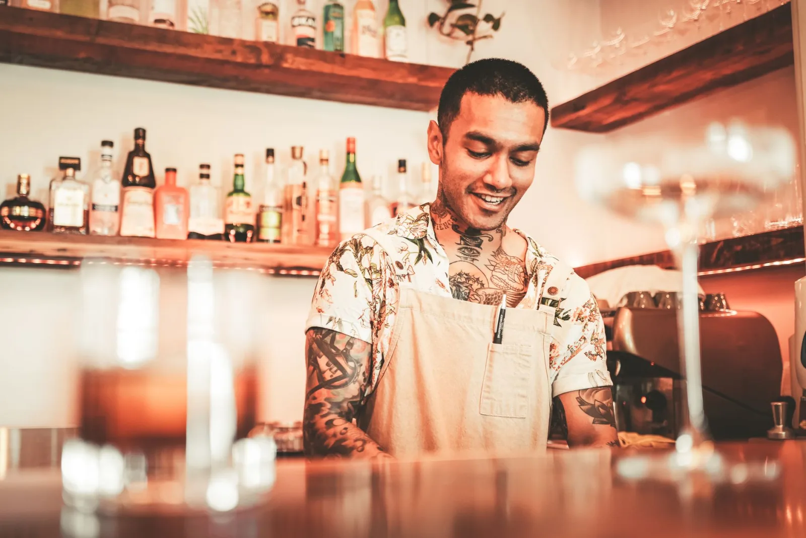 Tattooed bartender in apron preparing cocktails at a contemporary bar setting, surrounded by various liquor bottles.