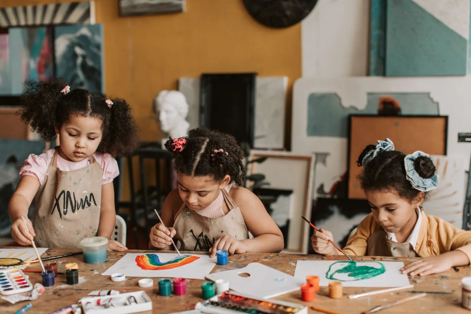 Three children painting creatively at a wooden table in an art class.