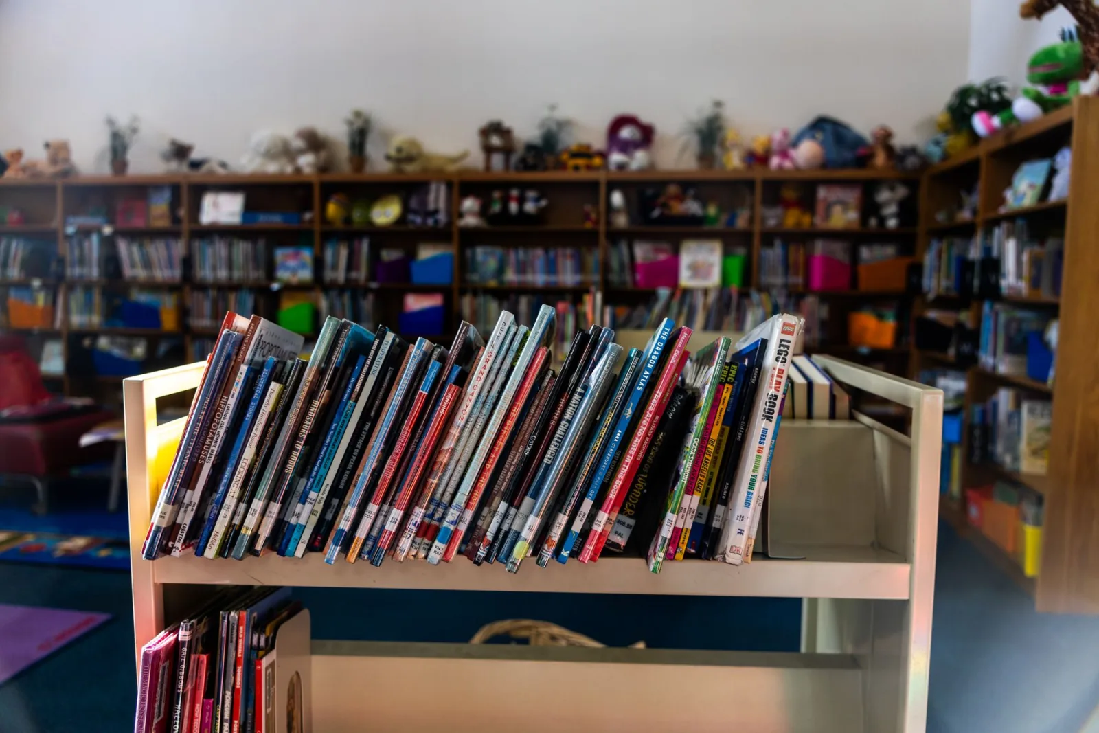 A warm library interior featuring a book cart in focus with colorful children's books on shelves.