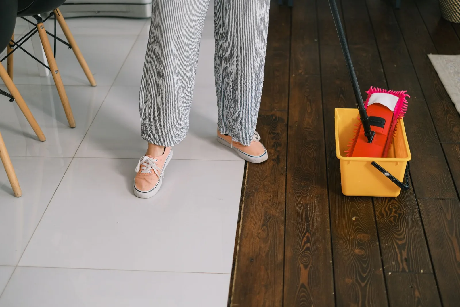 Crop anonymous woman in gumshoes and pants dipping mop into bucket with water on parquet in house