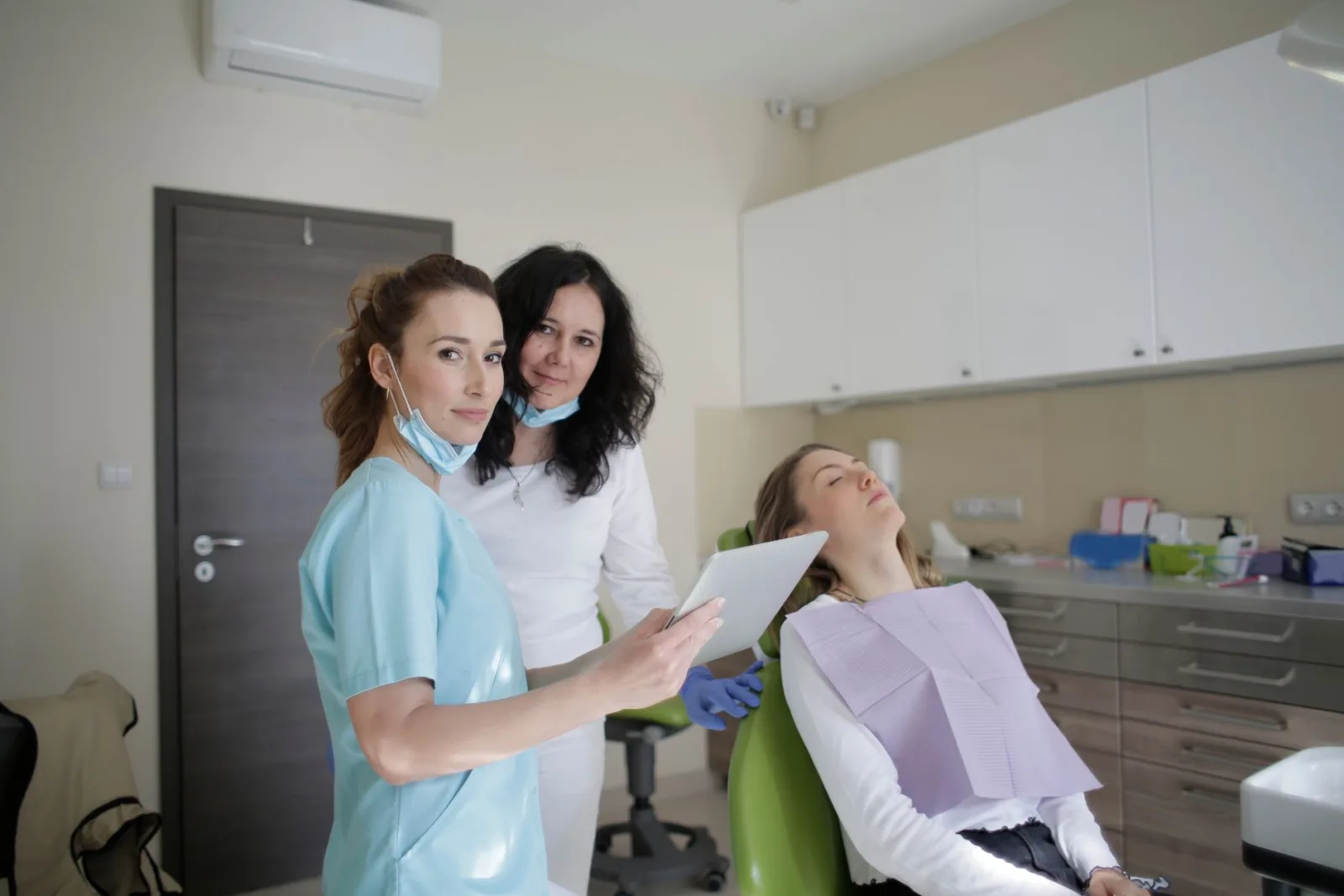 Smiling dentist in sterile mask and uniform looking at camera and showing tablet screen to cheerful female assistant while standing near patient with closed eye